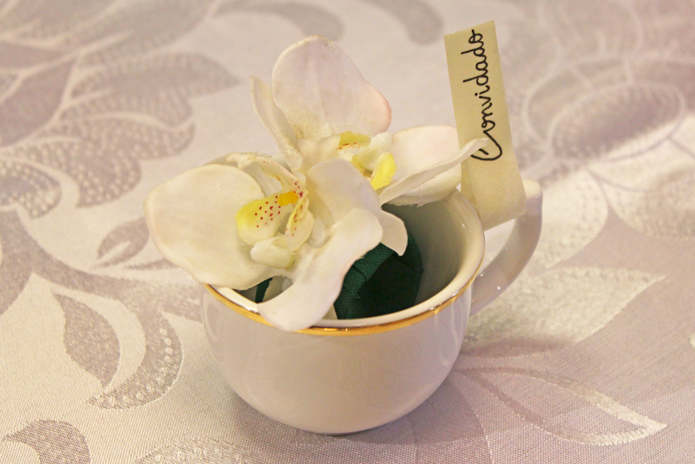 Imagem: Uma xícara de café da linha Flamingo Sofia, uma pequena flor e uma tira de papel. O truque é usar um papel mais consistente, com mais gramatura, e fita dupla face para unir os dois lados.