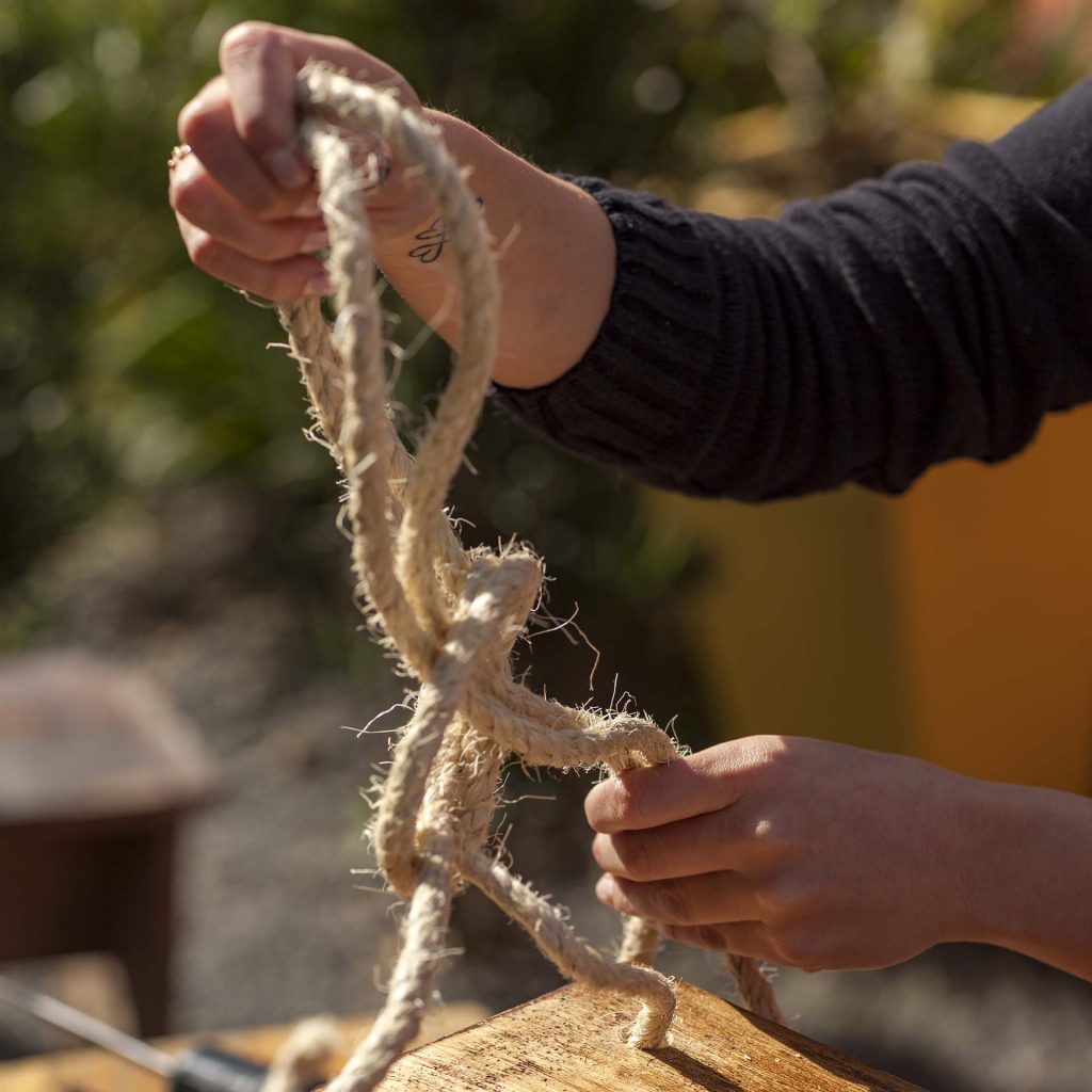 Imagem: Passo 3: Passe a corda pelos furos e dê um nó logo acima para unir as duas pontas. Confira se os nós estão na mesma altura para garantir o equilíbrio da peça. Foto: Raphael Günther/Bespoke Content.