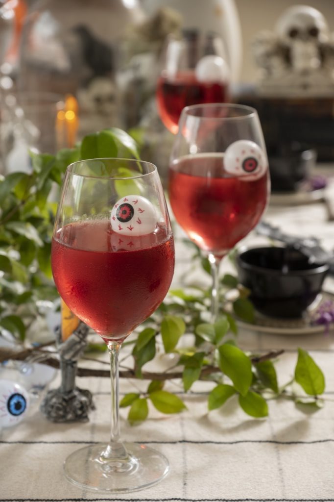 Foto aproximada da mesa posta Halloween mostra duas taças com líquido vermelho e um olho de plástico com íris vermelha, boiando em cada um.