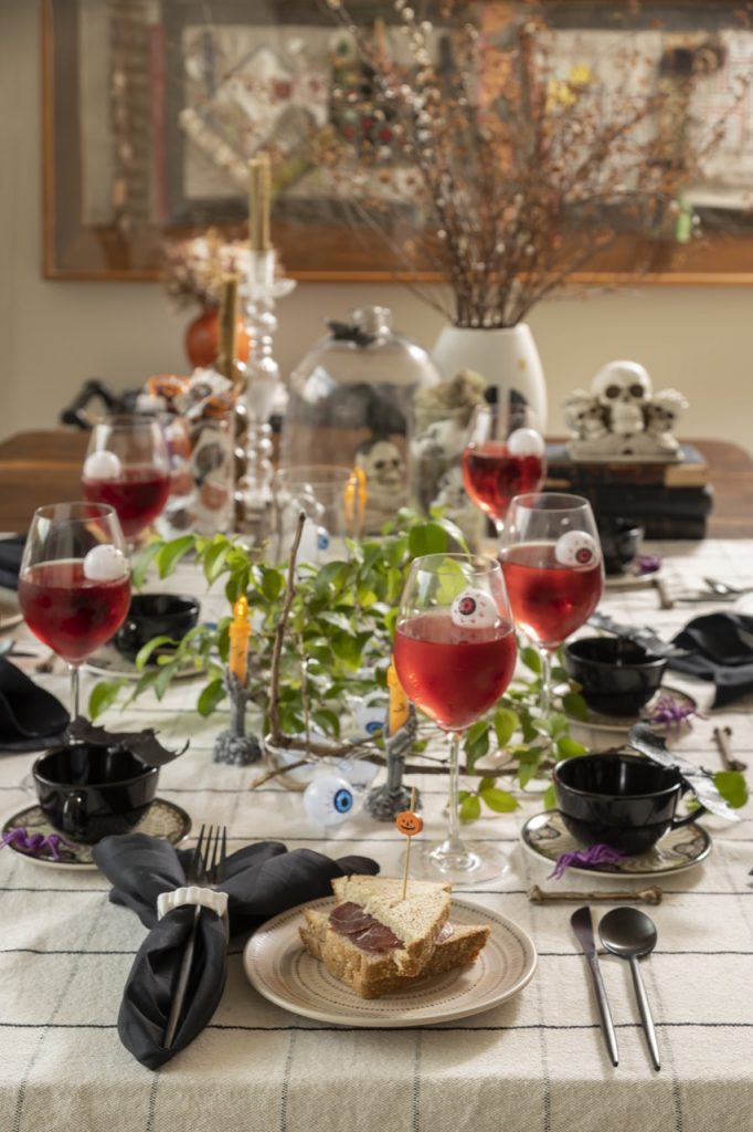 Mesa posta para o Halloween com pratos em bege e xícaras pretas. Há taças com líquido vermelho e olhos de plástico boiando. 