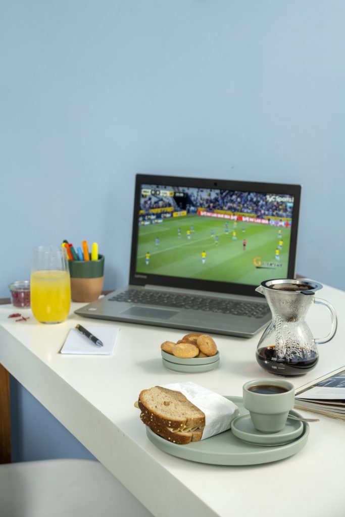 Sobre uma mesa branca, encostada a uma parede azul clara, temos: um notebook cinza com cena de jogo do Brasil na tela, um passador de café, xícara com cafezinho, prato com sanduíche, pires com biscoitos, caneta, bloco de papel, copo com suco e copo de cerâmica com canetas coloridas. 