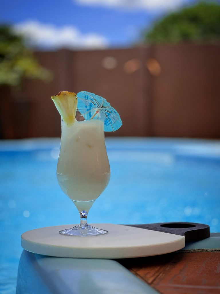 Imagem: pina colada servida em taça com guarda-chuva decorativo à beira da piscina. 