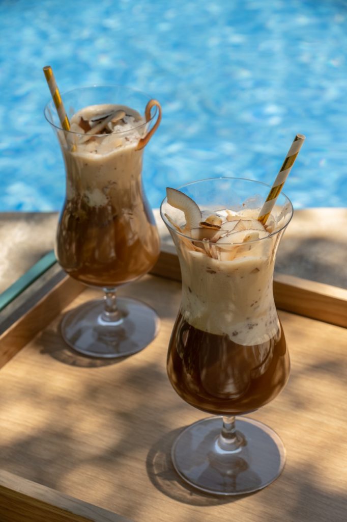 Sobre uma bandeja de madeira, vemos dois drinks gelados servido em taças com formato tulipa, com canudinhos de papel. 