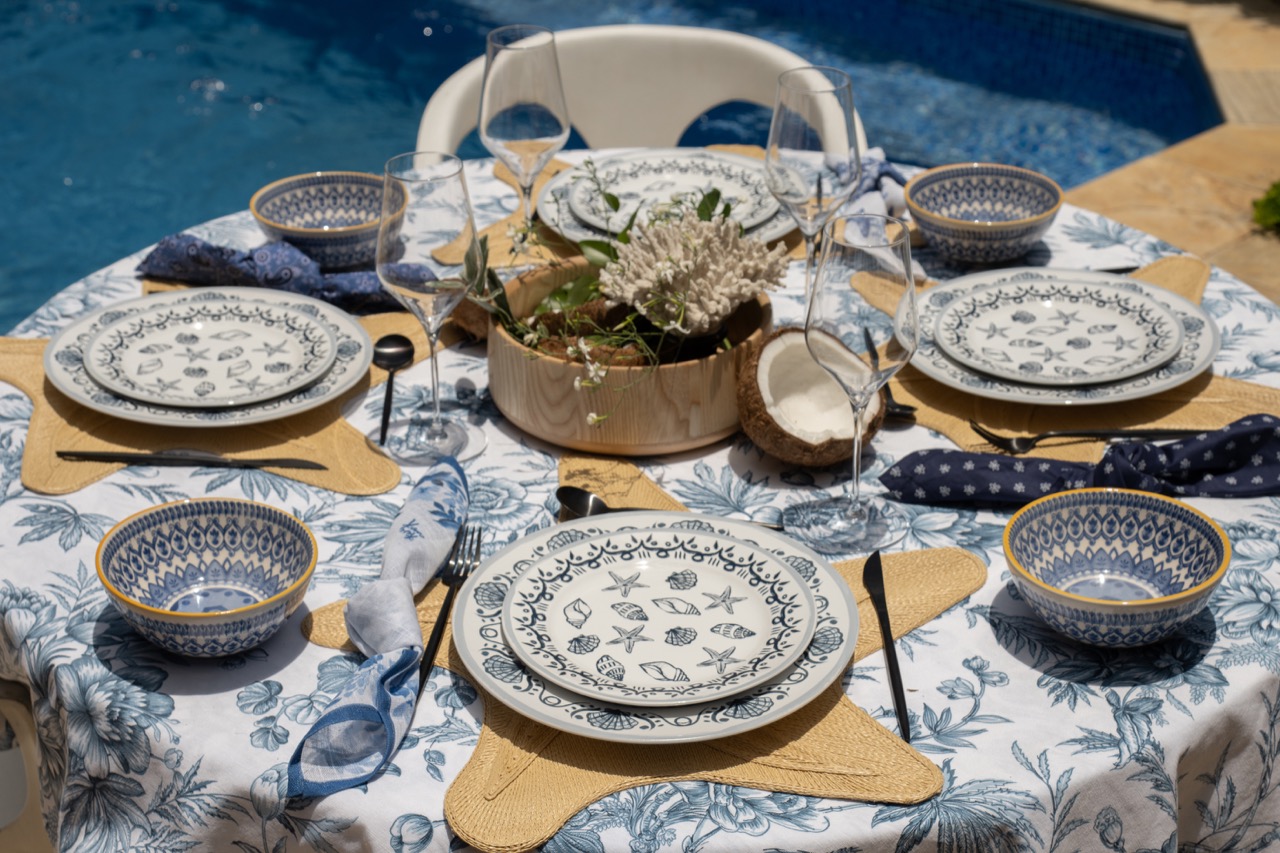 Mesa redonda, à beira da piscina. Cada lugar na mesa posta tem sobreposição de tigela, prato de sobremesa e prato raso. Tudo estampado em azul e branco, assim como a toalha. 

