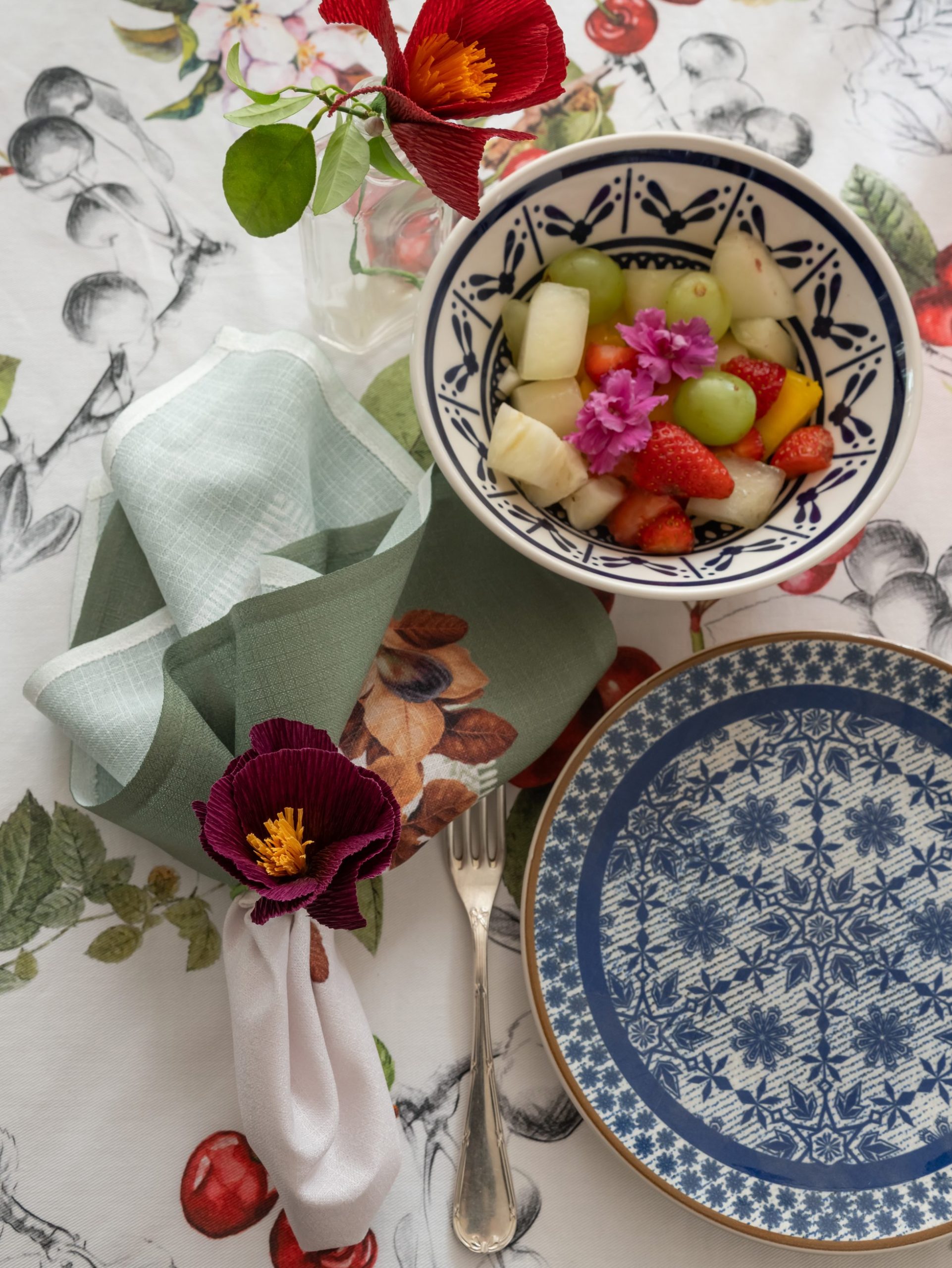 Imagem: mesa de café da manhã com louças azuis, bowl de frutas e toalha de mesa de cerejas.