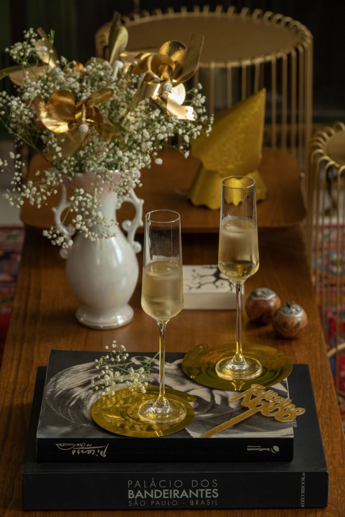 Duas taças com espumante estão sobre pratinhos dourados, apoiados sobre dois livros de arte, numa mesa lateral. 