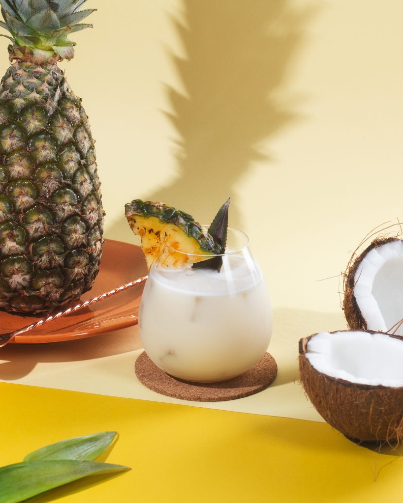 Imagem: copo de cristal da linha Easy da Oxford servido com pina colada de cor branca. Abacaxi e coco compõem a cena.