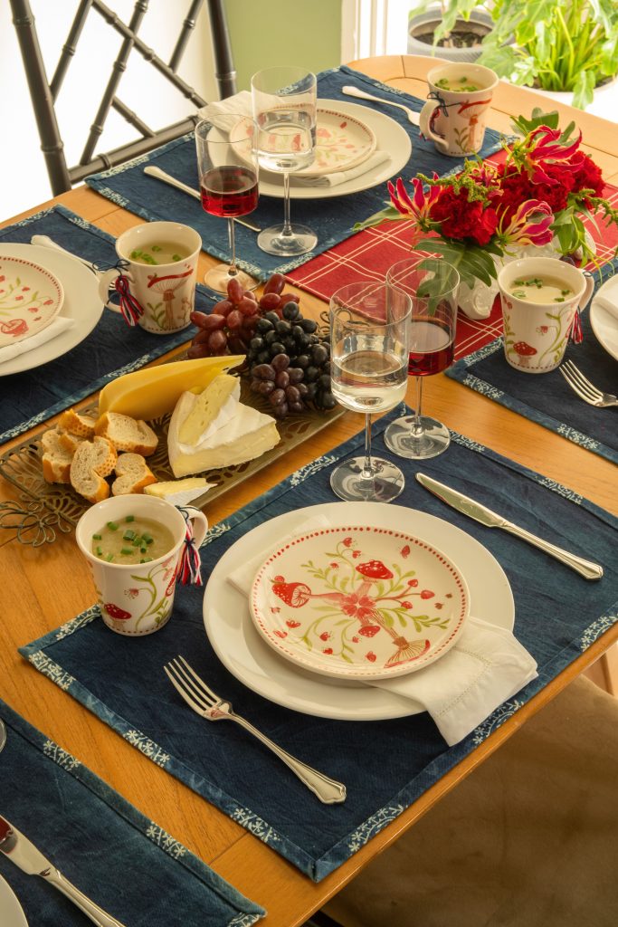 Imagem: mesa posta com caminhos de mesa vermelho e azul, prato de sobremesa e canecas de cogumelos. Taças de cristal servidas com água e vinho tinto.