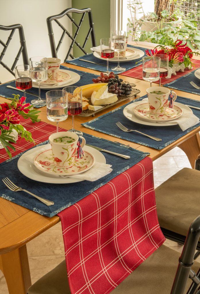 Imagem: mesa posta com caminhos de mesa vermelho e azul, prato de sobremesa e canecas de cogumelos. Taças de cristal servidas com água e vinho tinto.