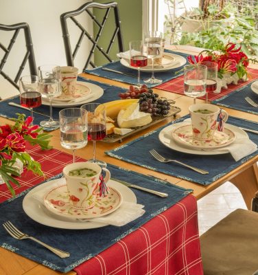Imagem: mesa posta com caminhos de mesa vermelho e azul, prato de sobremesa e canecas de cogumelos. Taças de cristal servidas com água e vinho tinto.