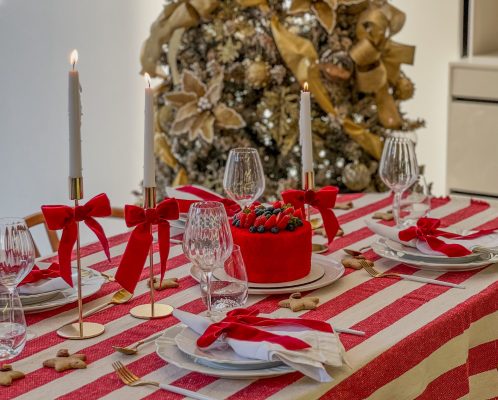 Imagem: mesa posta de natal com toalha listrada, pratos brancos e guardanapos com laços. Árvore de Natal ao fundo.