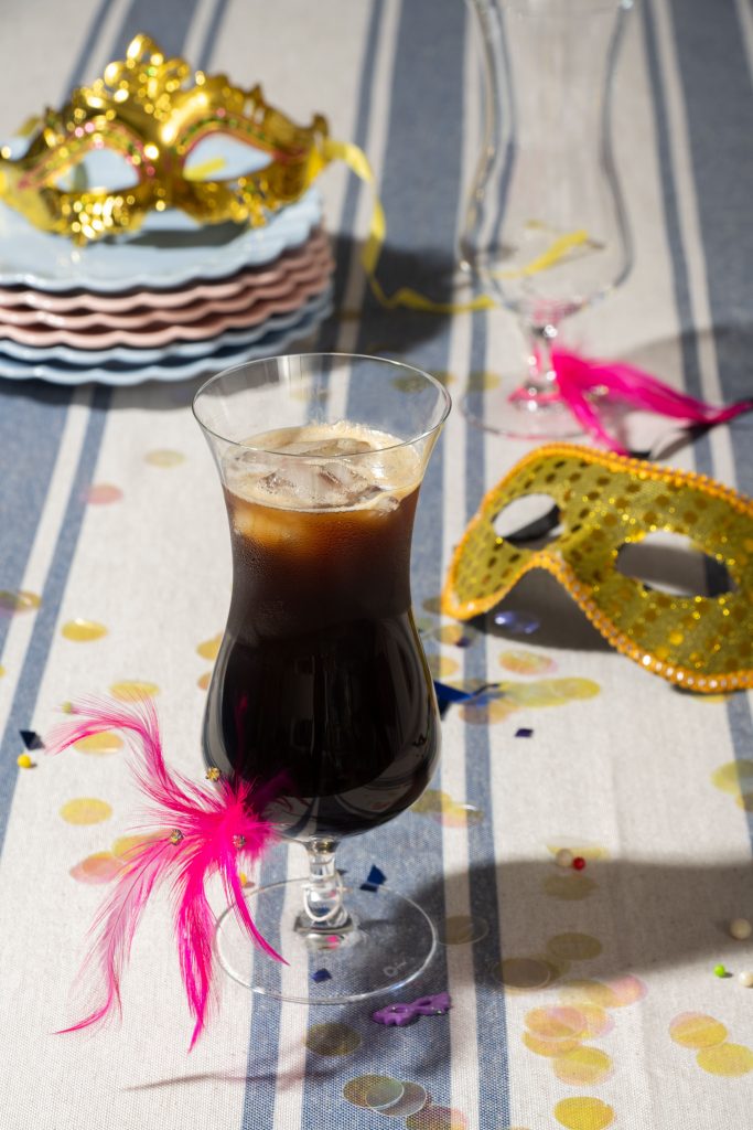 Imagem: Sobre a mesa, temos uma taça de cristal contendo drink gelado à base de café. Em segundo plano, vemos duas máscaras douradas de Carnaval, uma pilha de pratos em tom de azul ou rosa, lantejoulas e plumas em rosa pink.